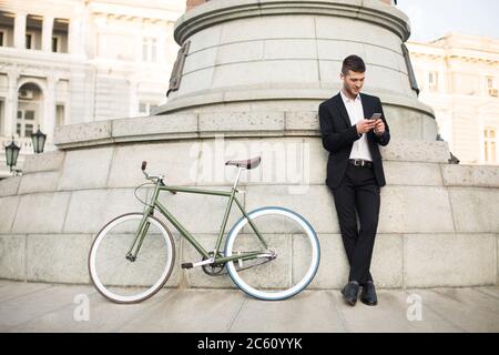 Young handsome man in classic black suit and white shirt with wireless earphones dreamily using cellphone with retro bicycle near on city street Stock Photo