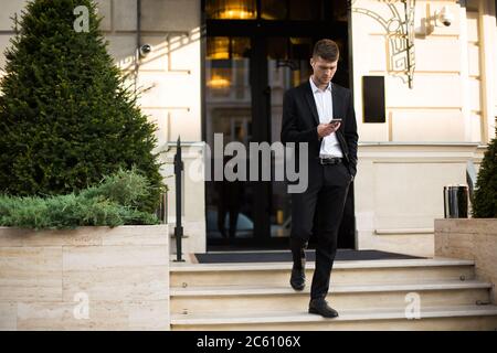 Young attractive businessman in black suit and white shirt with wireless earphones thoughtfully using cellphone while going down the stairs outdoor Stock Photo
