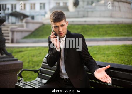 Young businessman in classic black jacket and white shirt indignantly looking aside while talking on cellphone on bench outdoor Stock Photo