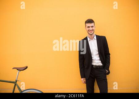 Young smiling man in classic black suit and white shirt happily looking in camera with bicycle near over orange background Stock Photo