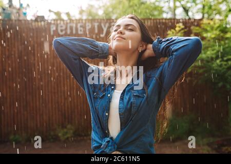 Portrait of young beautiful lady in denim shirt dreamily closing her eyes while standing under rain in park Stock Photo