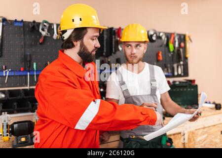 Two builders in work clothes and hardhats working in workshop wi Stock Photo
