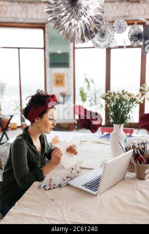 Young gorgeous woman with dark curly hair sitting at the table thoughtfully showing sketch in laptop while spending time in modern cozy workshop with Stock Photo
