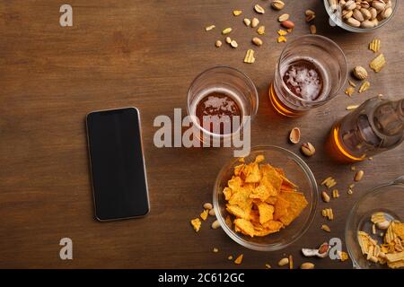 Modern get-togethers in bar. Smartphone, two glasses of beer, bottle, pistachio and chips are scattered on table Stock Photo