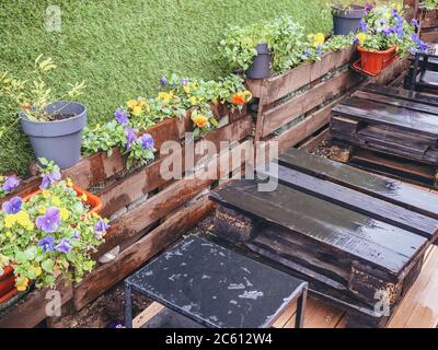 Wooden pallets wet from the rain stand near a wooden border with flowers next to a wall with a decorative green artificial grass coating Stock Photo