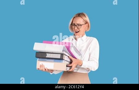 Stressed female secretary overloaded with paperwork, holding folders with documents on blue background Stock Photo