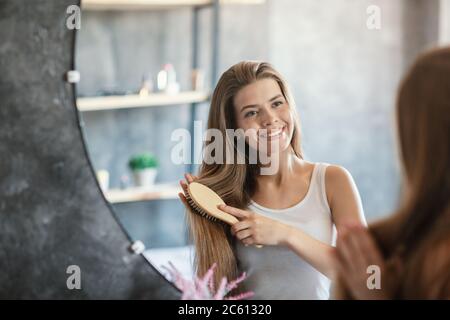 Lovely young girl brushing her beautiful long hair with wooden brush near mirror in bathroom Stock Photo