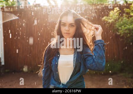 Portrait of beautiful lady in denim shirt dreamily looking in camera while standing under rain in park Stock Photo