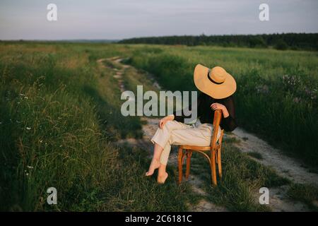 Slow living. Stylish elegant girl in straw hat sitting on rustic chair in summer meadow in evening. Fashionable young woman relaxing in field, tranqui Stock Photo