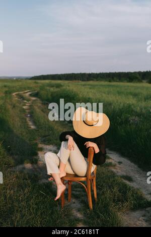 Stylish elegant girl in straw hat sitting on rustic chair in summer meadow in evening. Fashionable young woman relaxing in field, tranquil moment. Cre Stock Photo