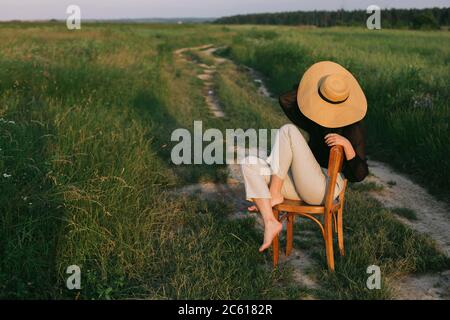 Fashionable young woman relaxing in field, tranquil moment. Creative image. Summer countryside. Stylish elegant girl in straw hat sitting on rustic ch Stock Photo