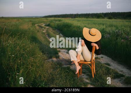 Stylish elegant girl in straw hat sitting on rustic chair in summer meadow in evening. Fashionable young woman relaxing in field, tranquil moment. Cre Stock Photo