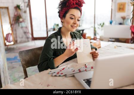 Young attractive woman with dark curly hair sitting at the table dreamily showing sketch in laptop while spending time in modern cozy workshop with Stock Photo