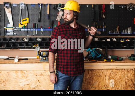 Builder in hardhat holding hammer on shoulder thoughtfully looki Stock Photo
