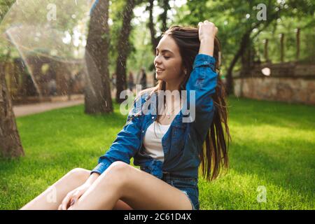 Young beautiful happy lady in shorts and denim shirt dreamily closing her eyes while sitting on grass under rain in park Stock Photo