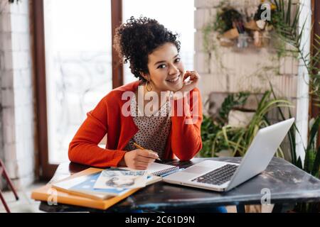 Young attractive smiling woman with dark curly hair sitting at the table happily working on laptop in modern workshop with big windows on background Stock Photo