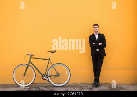 Young handsome man in classic black suit thoughtfully looking in camera with bicycle near over orange background Stock Photo