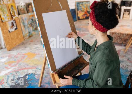 Young pretty woman with dark curly hair from back sitting on chair dreamily drawing picture on canvas while spending time in cozy art workshop Stock Photo