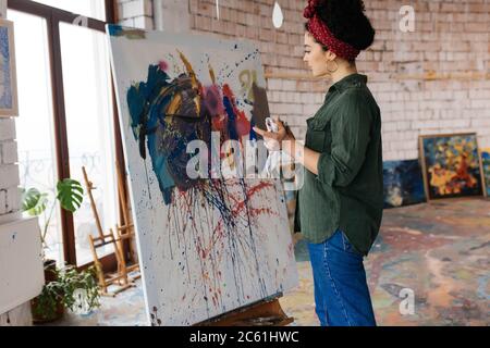 Young beautiful woman with dark curly hair thoughtfully drawing picture on canvas by hands with bright oil paints while spending time in big cozy Stock Photo