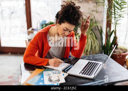 Young thoughtful woman with dark curly hair sitting at the table dreamily working on laptop in modern workshop with big windows on background Stock Photo