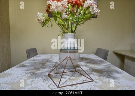 Copper pyramid, with Astromelia vase, Alstroemeria Hybrida on a travertine marble table base with white lace tablecloth, with chairs and white wall in Stock Photo