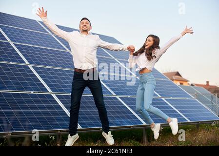 Young cheerful man and woman jumping holding hands by a solar panel, wearing similar clothes, concept of happy life Stock Photo
