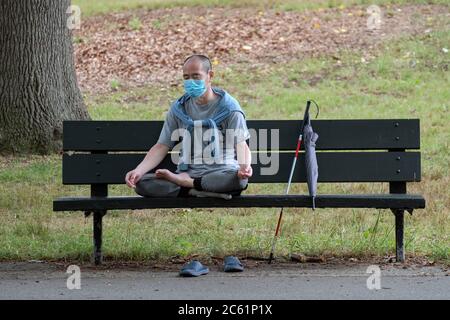 An apparently blind or visually impaired Asian American man wearing a surgical mask sits on a park bench and meditates. In Kissena Park, Flushing, NY. Stock Photo