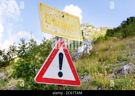 Yellow village traffic sign and triangular traffic sign with an exclamation mark  with bullet holes against blue sky, Montenegro Stock Photo