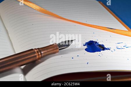A copper fountain pen splats a blue ink mess on a notebook with orange bindings and ribbon Stock Photo