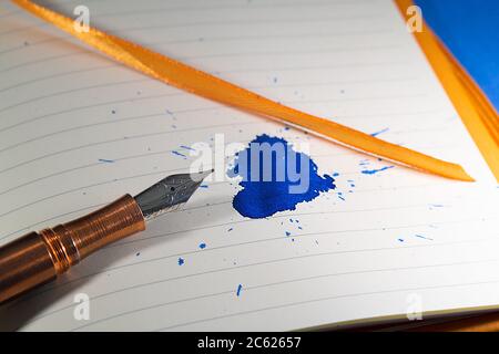 A copper fountain pen splats a blue ink mess on a notebook with orange bindings and ribbon Stock Photo