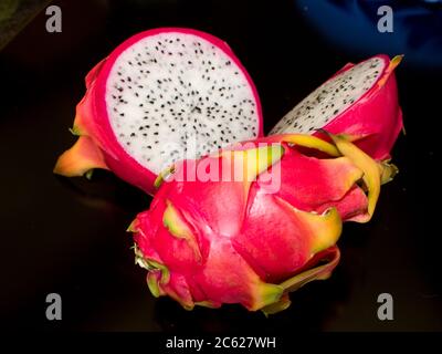 A Dragon Fruit, also known as a Pitaya, with a sliced dragon fruit, exposing the white flesh and small edible seeds, against a black background Stock Photo