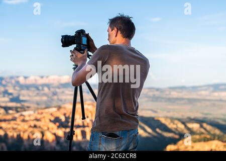 Man taking pictures photos of view from Bryce Point overlook of hoodoos rock formations in Bryce Canyon National Park at sunset with tripod and camera Stock Photo