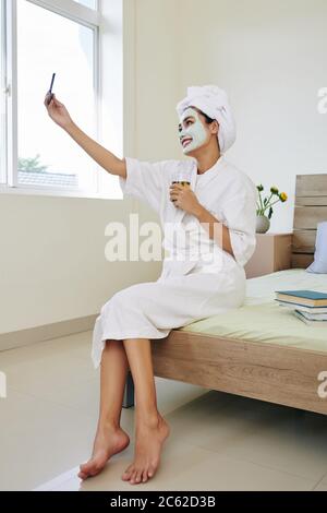 Happy beautiful young Asian woman in bath robe sitting on edge of her bed with retexturizing mask on her face drinking glass of green tea and taking s Stock Photo