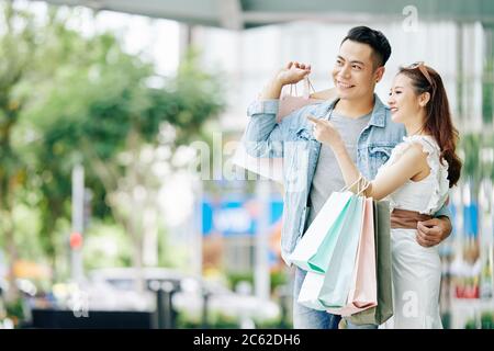 Beautiful happy young Asian couple standing in the street with shopping bags and looking away Stock Photo