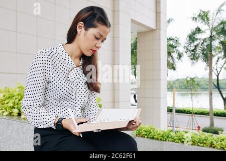 Serious young mixed-race businesswoman sitting outdoors and taking notes in her planner Stock Photo