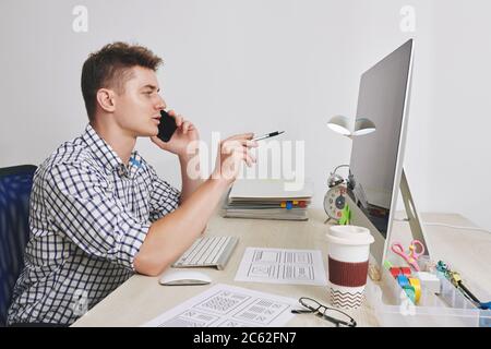 UX designer talking on phone with client and pointing at computer screen with pen Stock Photo