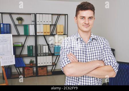 Portrait of smiling confident young UX designer standing in office with arms crossed and looking at camera Stock Photo