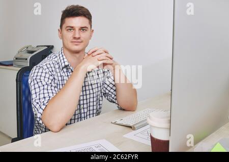 Portrait of handsome smiling young entrepreneur sitting at office desk and looking at camera Stock Photo