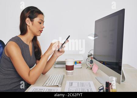 Young female UX designer working on project at her desk and reading text messages and e-mails from client on smartphone screen Stock Photo
