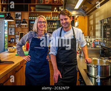 Chef Felix Groß in the open kitchen of Ernerle with food journalist Angela Berg. In the background, shelves full of regional specialties in Hittisau, Austria