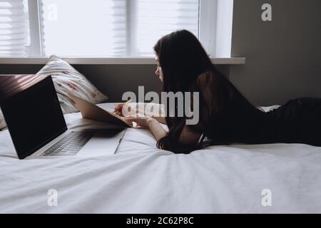 Young woman lies on bed with laptop and notebook. Stock Photo