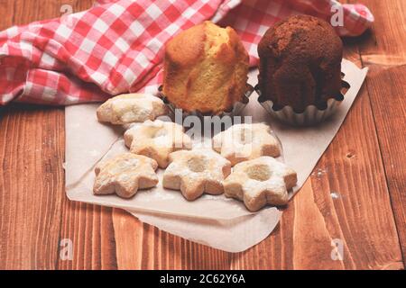 Tea cookies and muffins on wooden background. Homemade pastry concept. Star shaped biscuits lying on white cloth by red plaid kitchen towel. Composition of tiny gateau with sugar powder and cupcakes Stock Photo