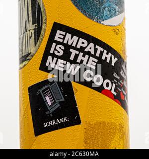 A black sticker reading "Empathy is the new cool" is stuck on a yellow lamp post along the boardwalk pier of Sankt Peter-Ording, Germany. Directly below it, a second sticker shows a wardrobe labeled "Schrank" (cabinet), with red hearts and the silhouette of a ship visible to the right. Stock Photo