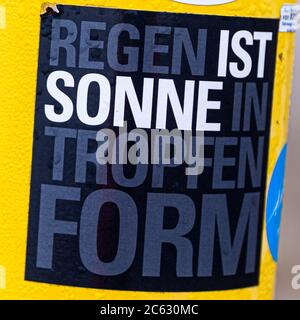 A black sticker reading "Regen ist Sonne in Tropfen Form" (rain is sun in the shape of drops) is stuck on a yellow lamp post along the boardwalk pier of Sankt Peter-Ording, Germany. Droplets on the painted surface show that it has actually been raining. Stock Photo