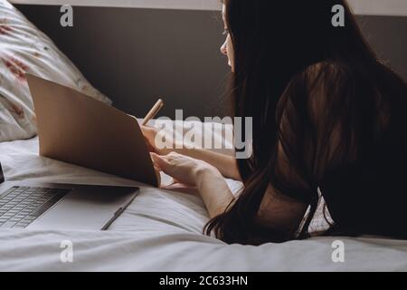 Young woman lies on bed with laptop and notebook. Stock Photo