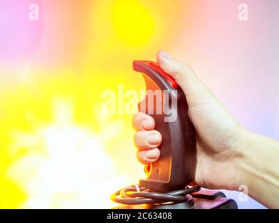 A hand holds a joystick for games by pressing the red button with a thumb on a multicolor background similar to an explosion Stock Photo