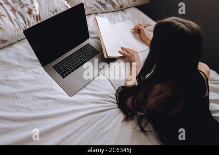 Young woman lies on bed with laptop and notebook. Stock Photo