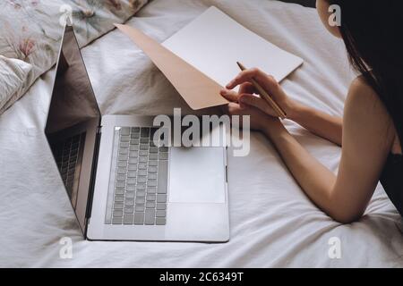 Young woman lies on bed with laptop and notebook. Stock Photo