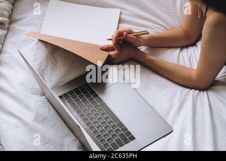 Young woman lies on bed with laptop and notebook. Stock Photo