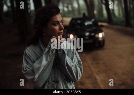 Close up of young woman standing alone in forest with car behind Stock Photo